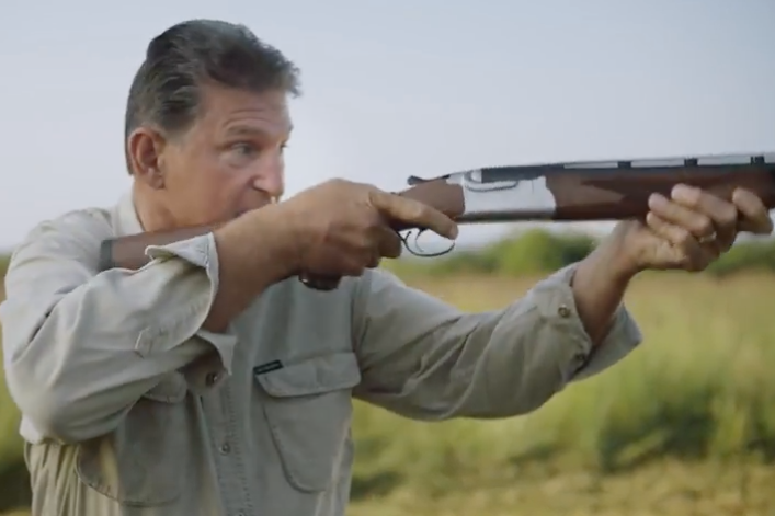 Manchin, standing in a field, levels and aims a shotgun.